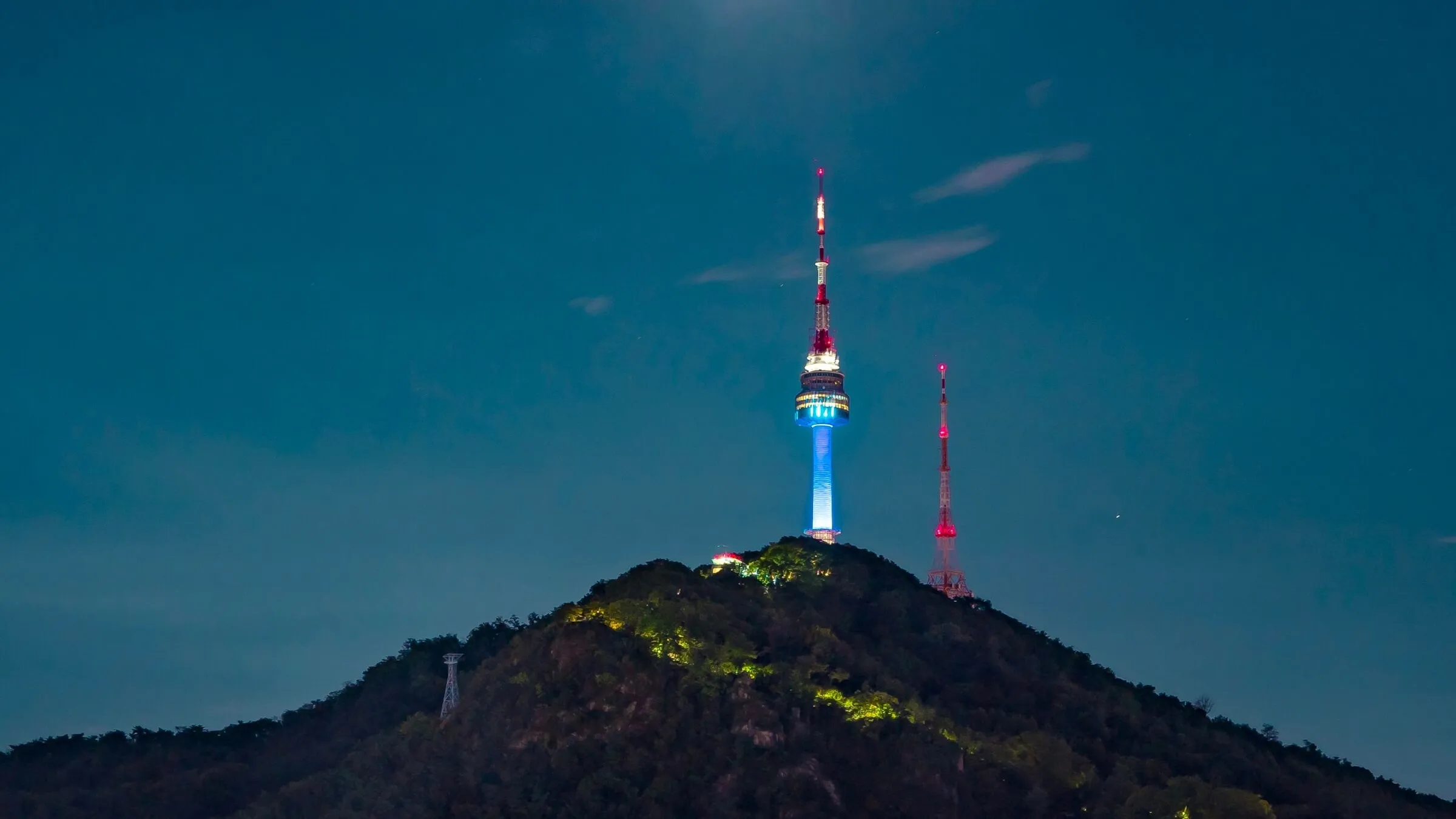 N Seoul Tower lit up in blue and red at night on top of the dark, wooded summit of Namsan, a broadcast mast beside it and a cable car pylon on the slope
